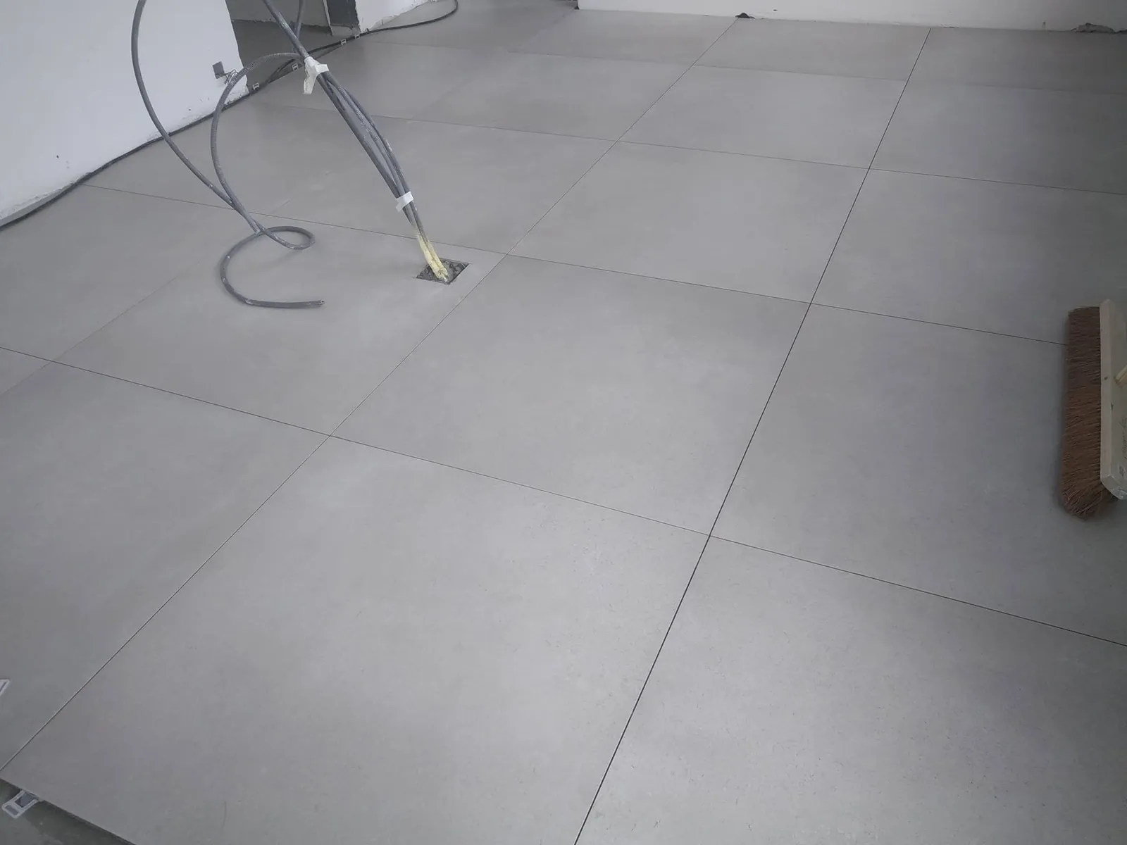 Gros plan sur de grands carreaux gris posés, gaine électrique sortant du sol et balai en bout de pièce