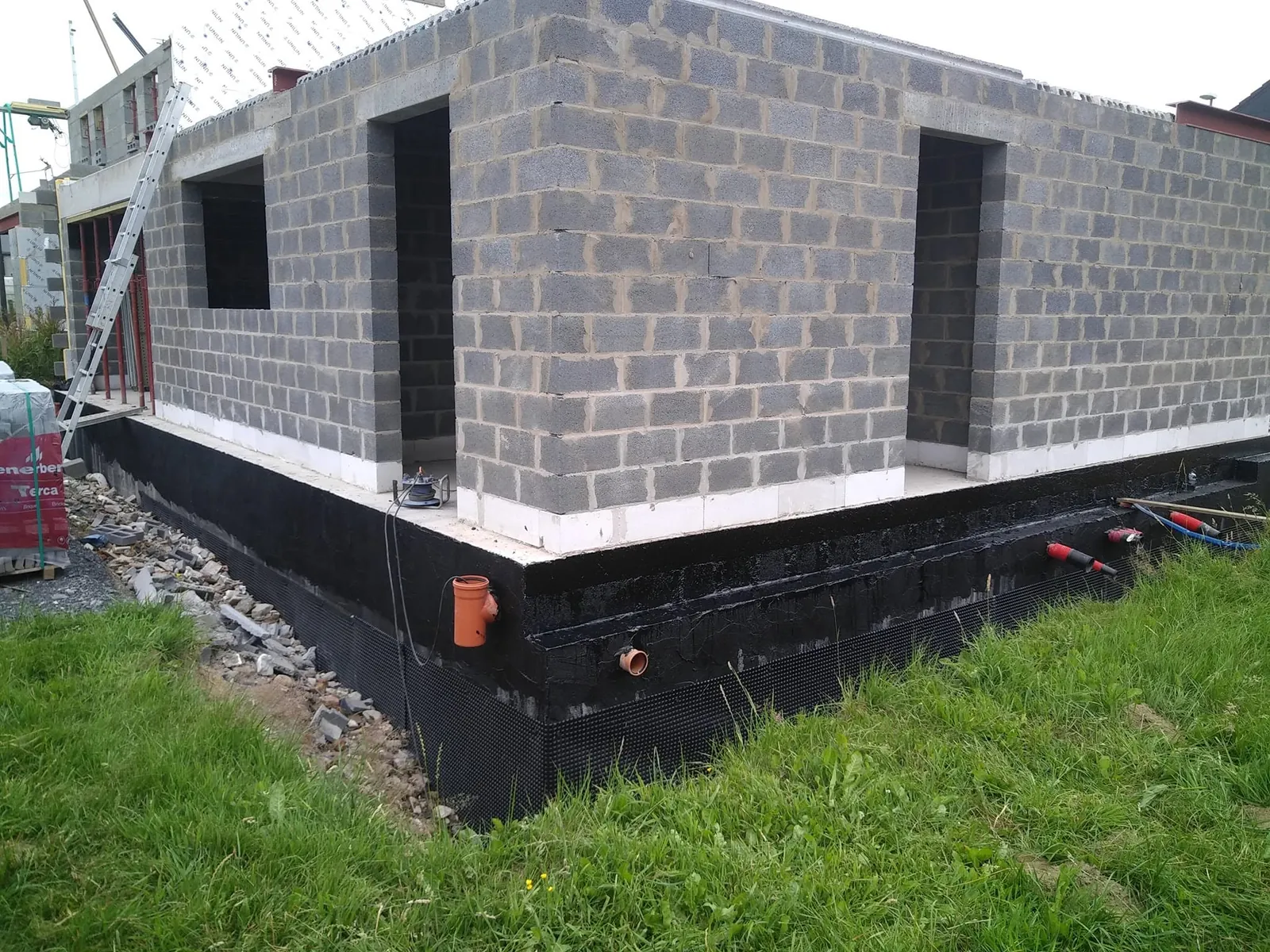 Angle de maison en blocs sur un soubassement recouvert d'une membrane noire d'étanchéité, tuyaux orange et gravier de drainage