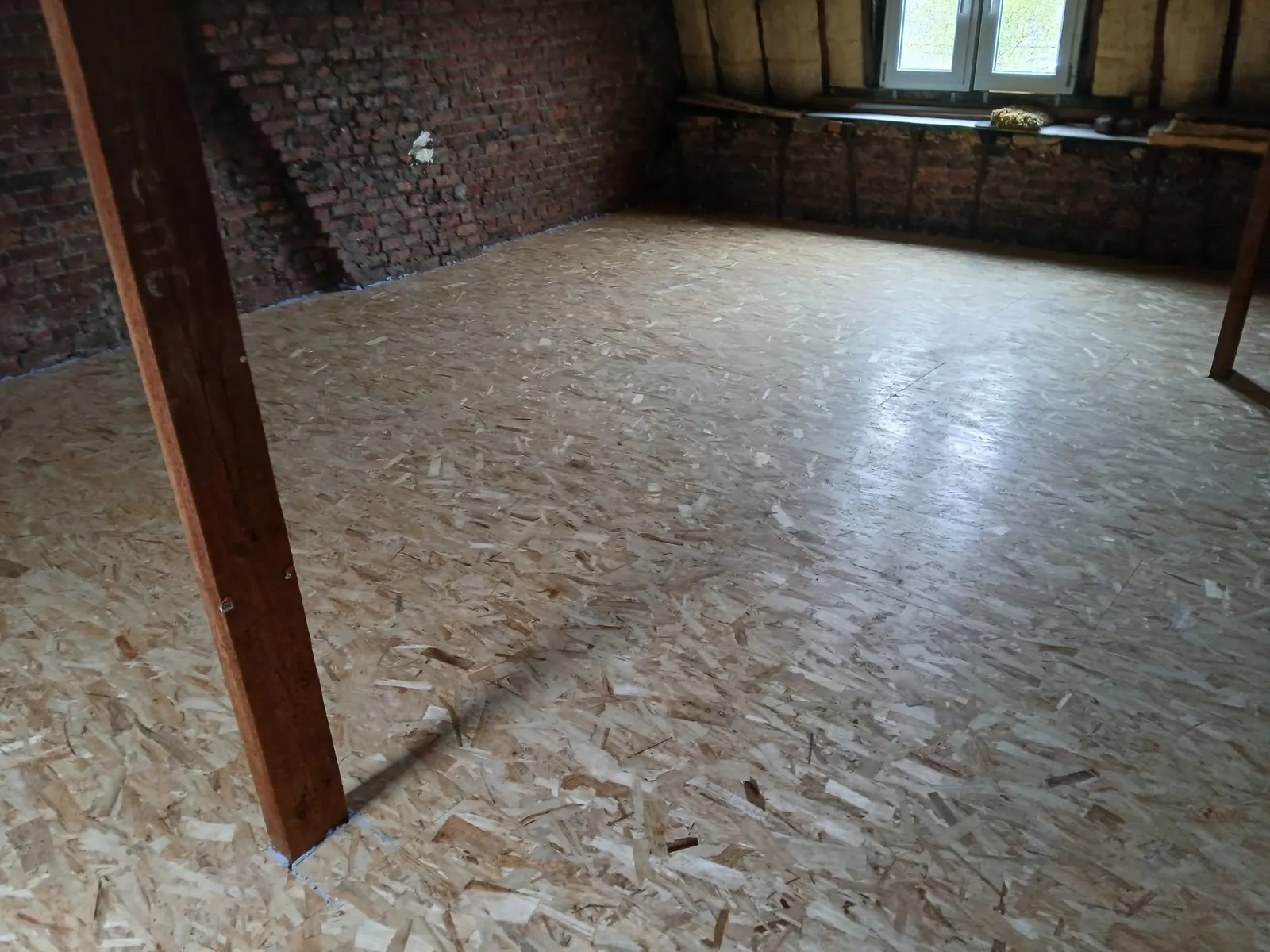 Plancher de combles en panneaux OSB, poutre en bois au premier plan, murs en briques et fenêtre au fond