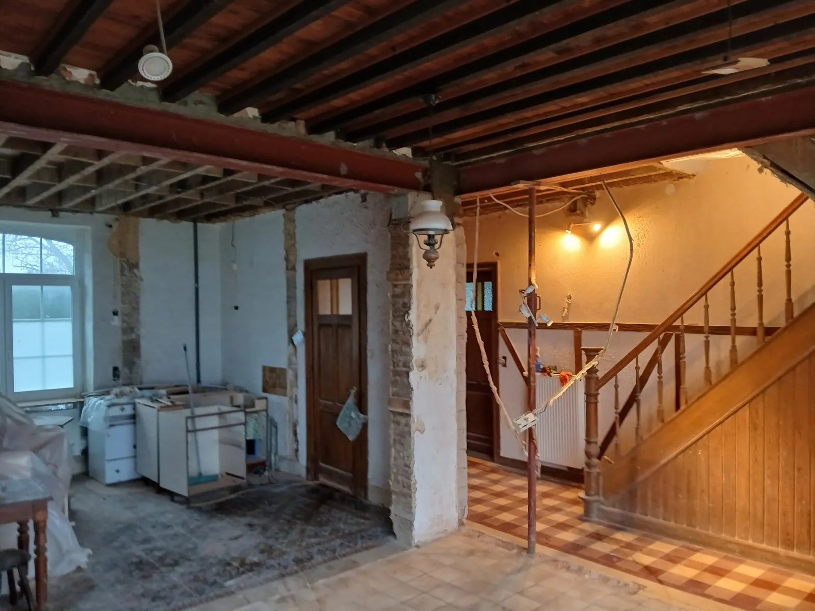 Pièce en démolition : poutre en acier sous un plafond à solives de bois, étai métallique et escalier en bois au fond