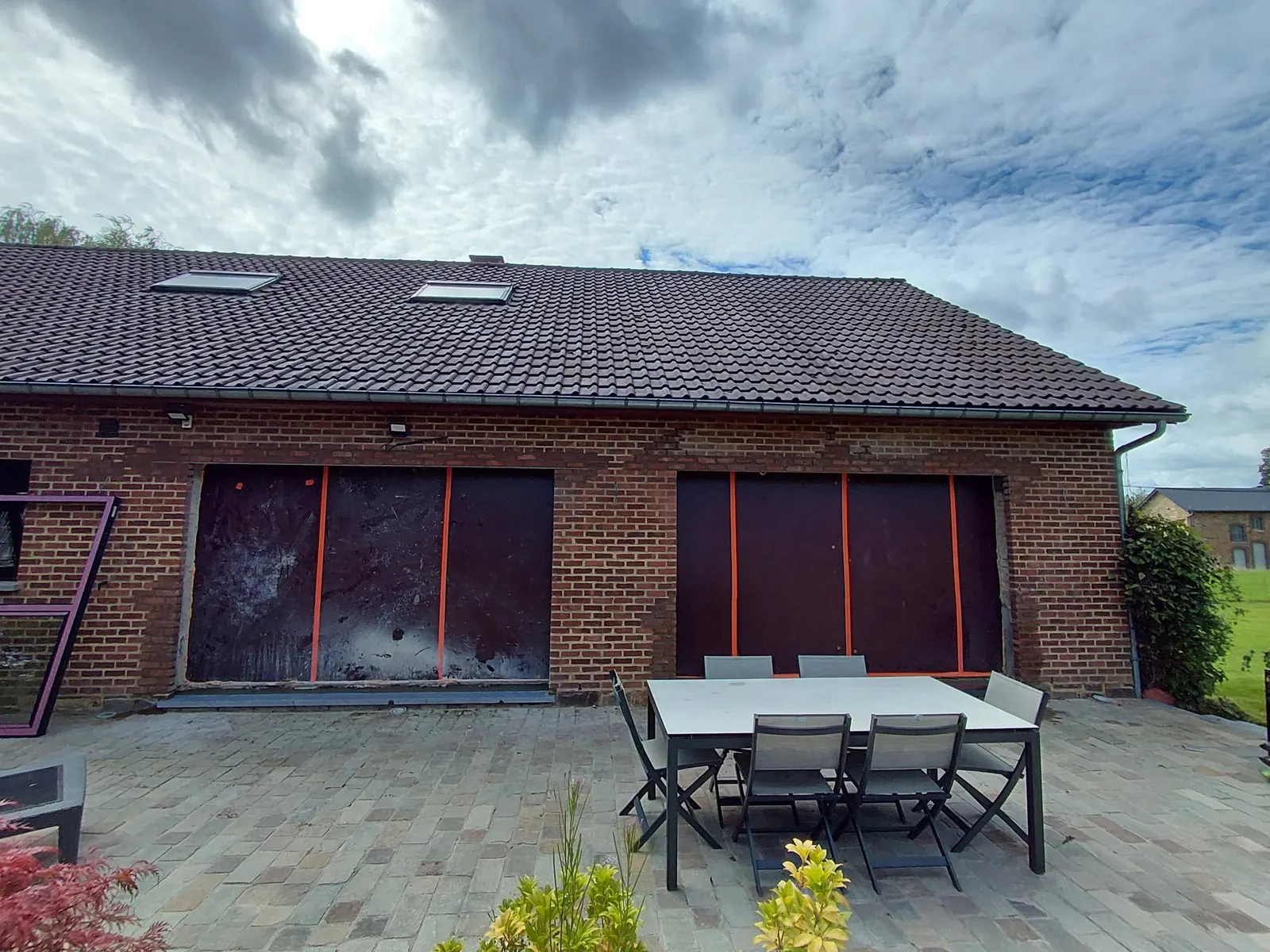 Deux grandes ouvertures fermées par des panneaux rouges dans une façade en briques, terrasse en pavés avec table et chaises