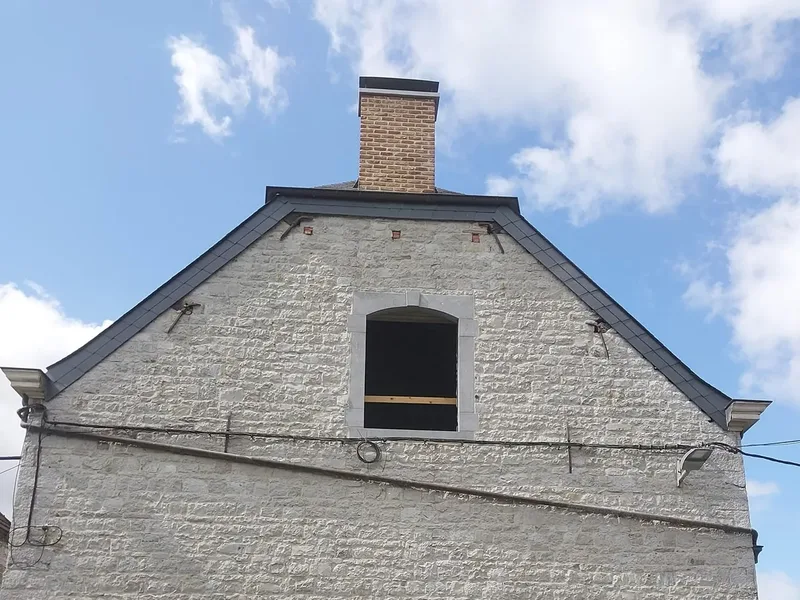 Pignon en pierre avec une petite fenêtre cintrée et une cheminée, haie au pied du mur