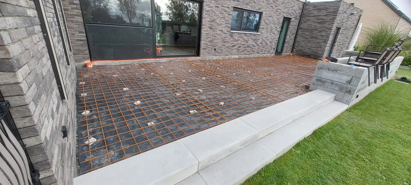 Treillis d'armature posé pour une terrasse contre une façade en briques grises, avec une marche en béton et la pelouse